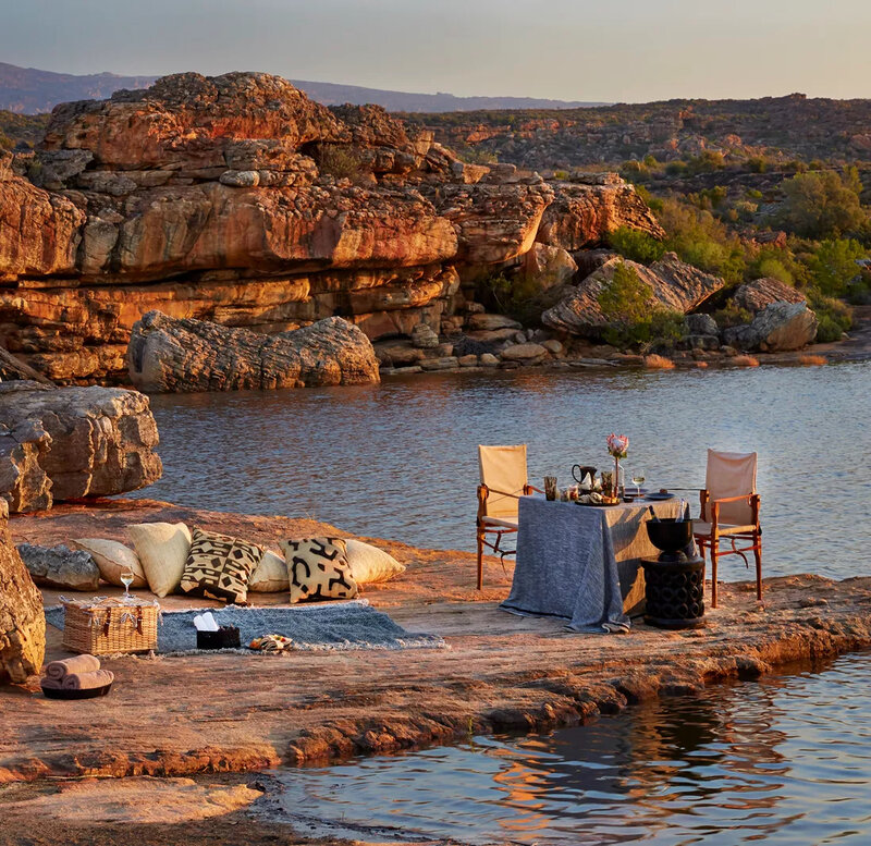 A romantic riverside dining setup in the South African wilderness, featuring elegant seating, plush cushions, and a beautifully set table at sunset.
