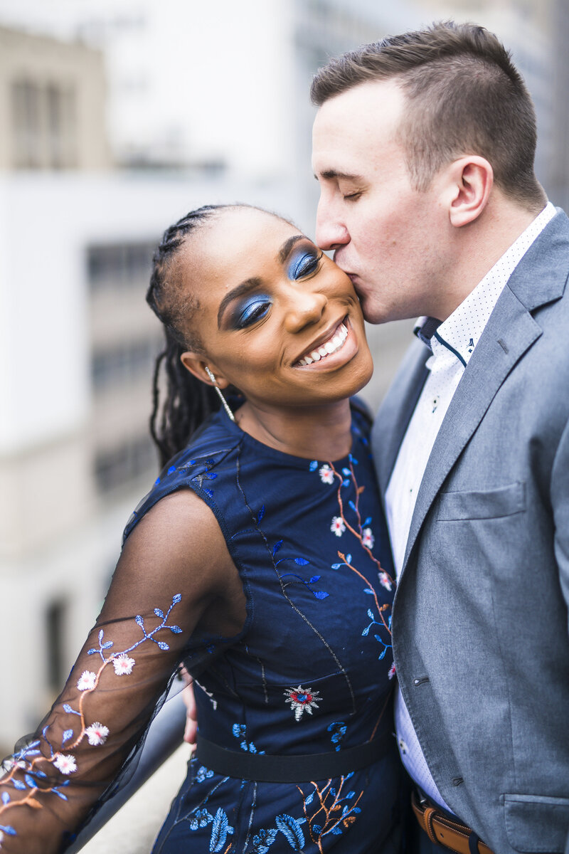 Fiance kisses his fiancee on the cheek on a rooftop photoshoot.