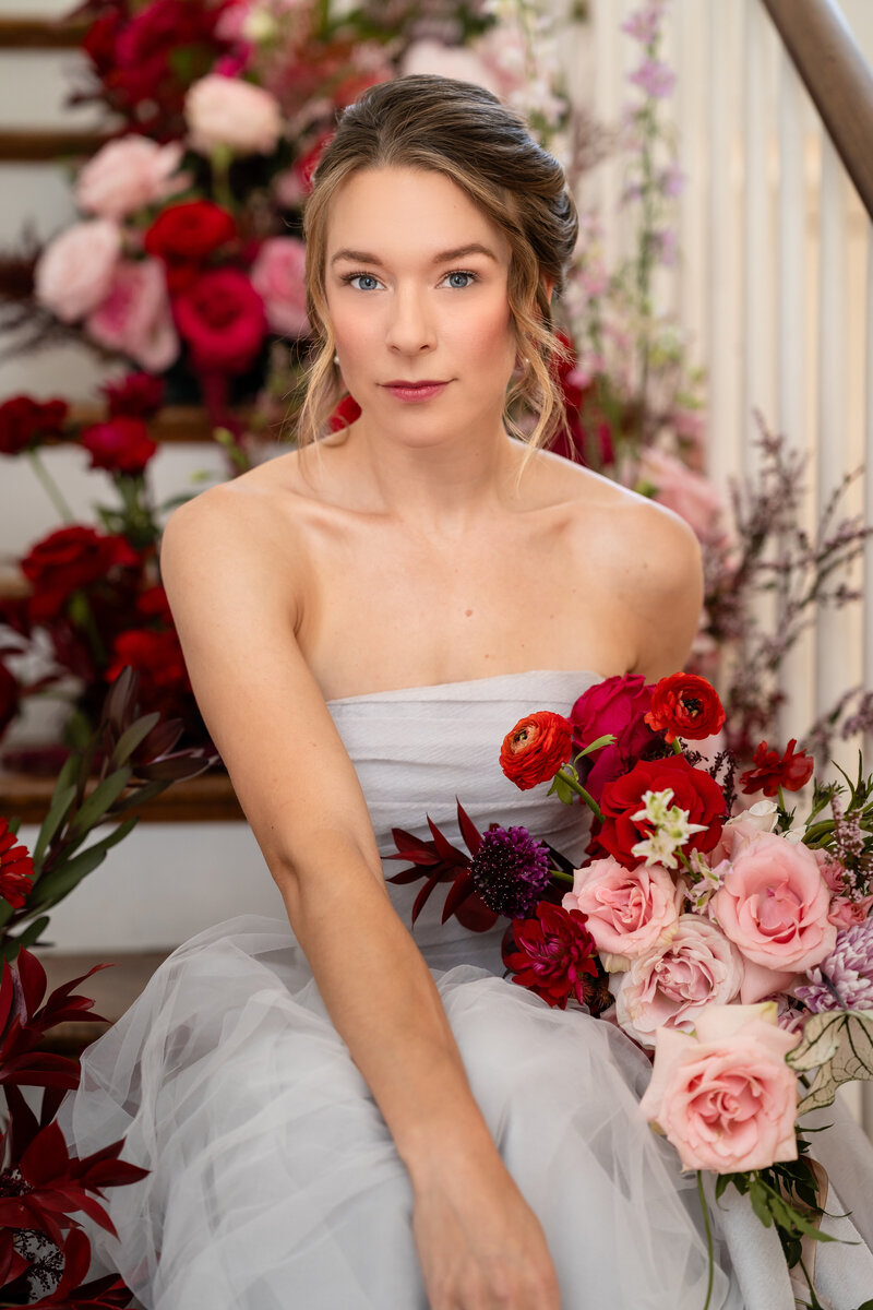 Bride sits on the stairs at Rust Manor House in Leesburg Virginia surrounded by pink and red flowers