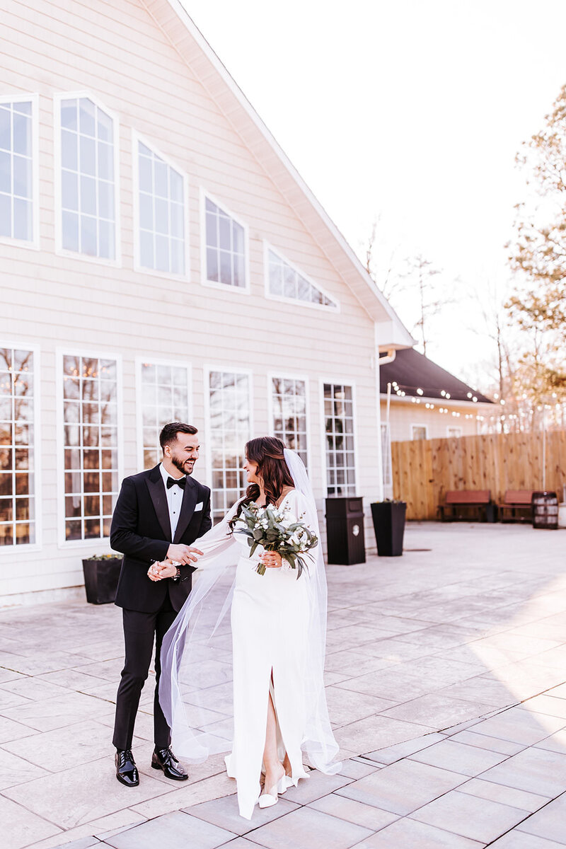 Groom in black tuxedo holds onto the arm of his bride, while they both run and laugh together, in front of the ballroom windows at Running Deer Golf Club.