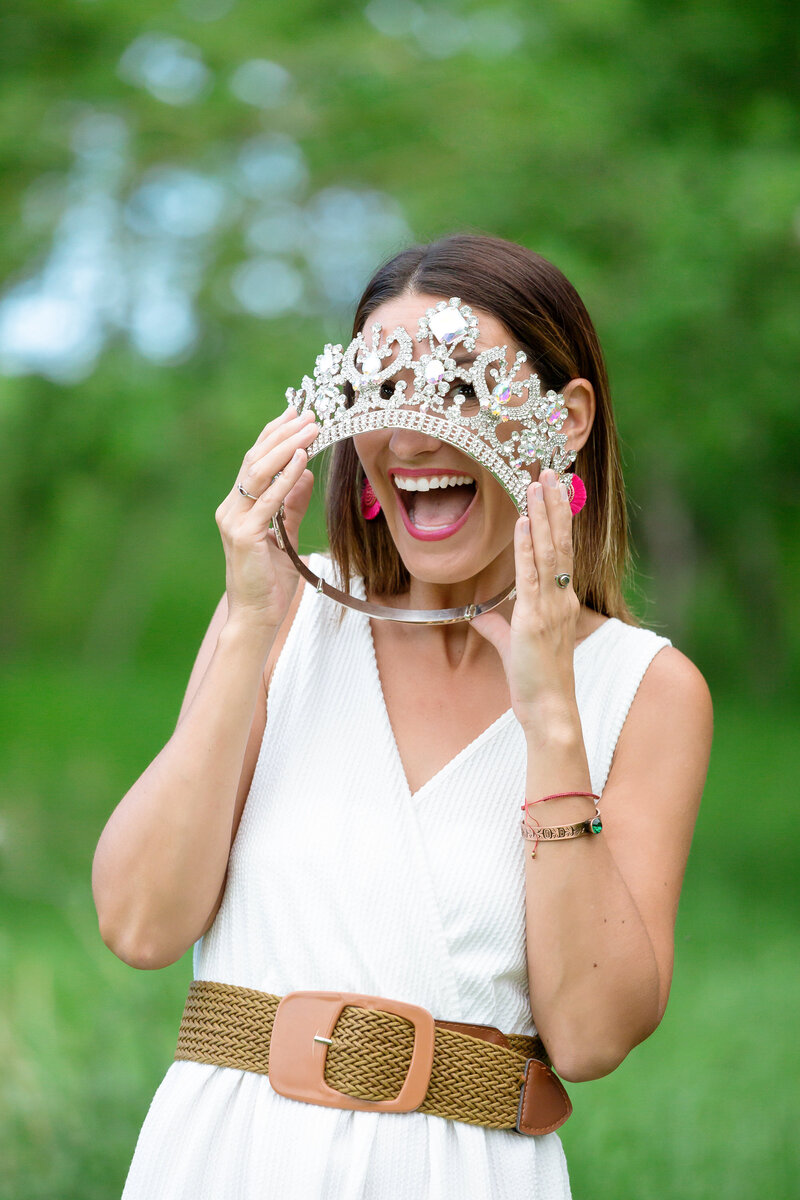 Calgary brand storytelling photoshoot of a woman playfully holding a sparkling jewelled crown in front of her face, framing her wide smile. She wears a white sleeveless dress cinched with a woven belt and accessorizes with pink earrings, bracelets, and rings. The lush green outdoor background adds a fresh and vibrant feel to the fun and confident portrait.