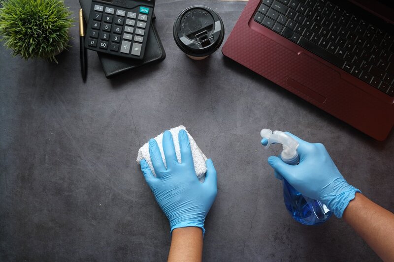 Paire de main avec des gants bleus, un spray dans la main droite et un chiffon dans la gauche pour nettoyer un bureau