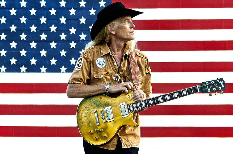 Country Music Photo Billy Crain standing in front of American Flag wearing black cowboy hat hands resting on guitar slung from shoulder