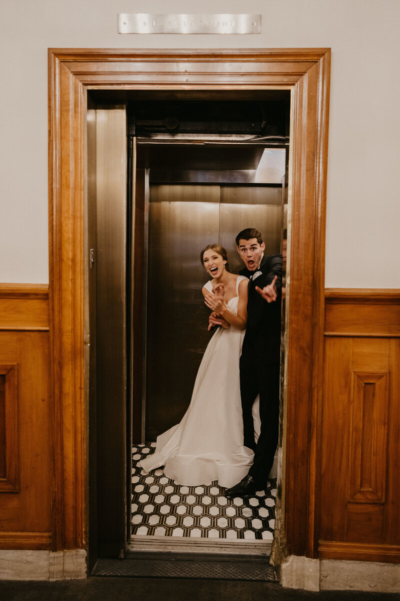 A bride and groom dressed in their wedding day attire laugh at the camera while riding an elevator because they were just caught kissing when the elevator doors opened
