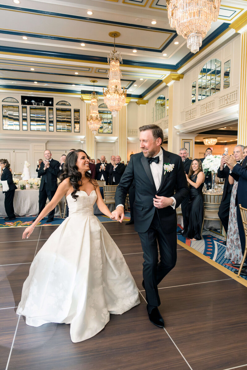 A bride and groom dance joyfully in an elegant ballroom, surrounded by guests clapping and smiling. The bride wears a strapless white gown, and the groom is in a black tuxedo. Crystal chandeliers and ornate decor adorn the background.