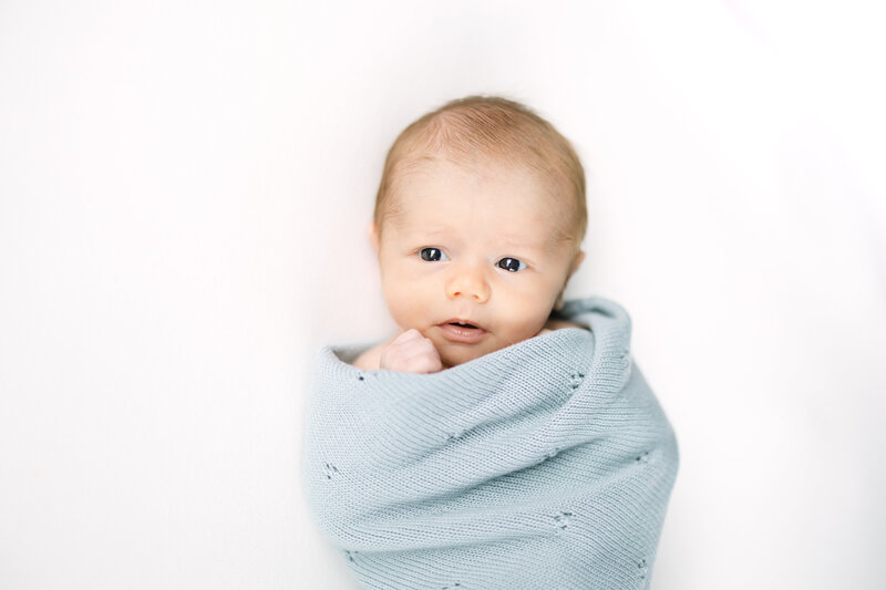 a baby boy lays on a white background, wrapped in a light blue baby blanket.