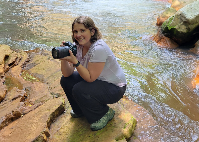 Joy Granados crouching on river rocks with her camera during an outdoor photoshoot, behind-the-scenes image of Capture Joy Photography, Northern Virginia family photographer.