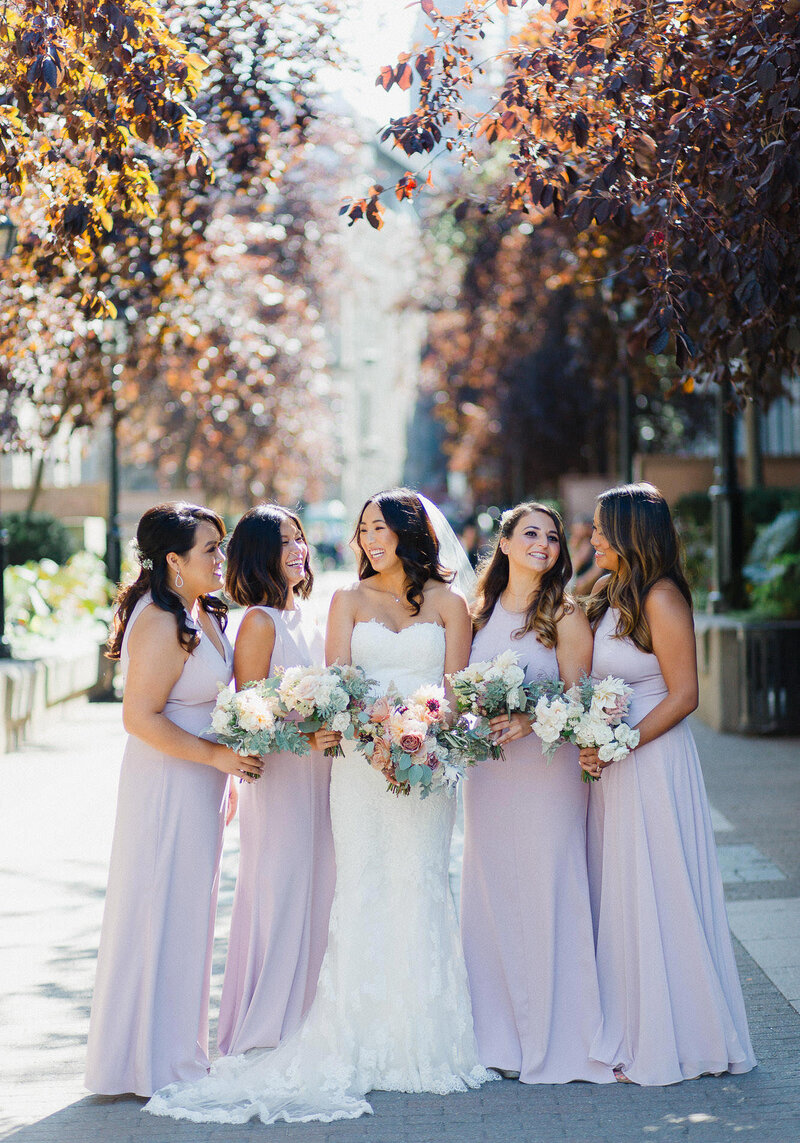 Demoiselles d'honneur avec la mariée, se regardent en riant, elles portent toute une magnifique robe mauve pâle et il y a des arbres avec des feuilles d'automne derrière. Photo de mariage