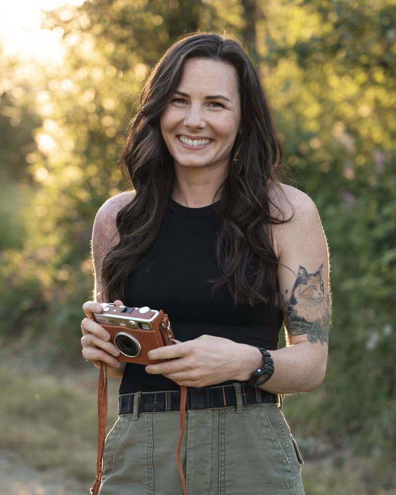 a female photographer smiles at the camera while holding a camera in her hands during golden hour