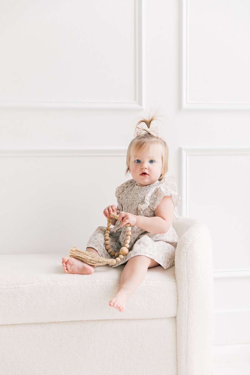 A baby girl sitting on a white couch holding a wooden toy during studio milestone session with  Charlotte baby photographer.