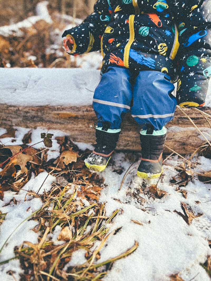 A child sitting on a log outdoors in winter, wearing colorful space-themed clothing and snow boots, surrounded by snow and dried grass.