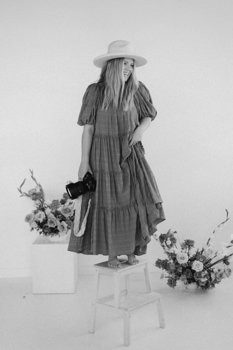 Black and white photo of a photographer standing on a stool surrounded by floral arrangements, capturing an artistic and dynamic branding moment.