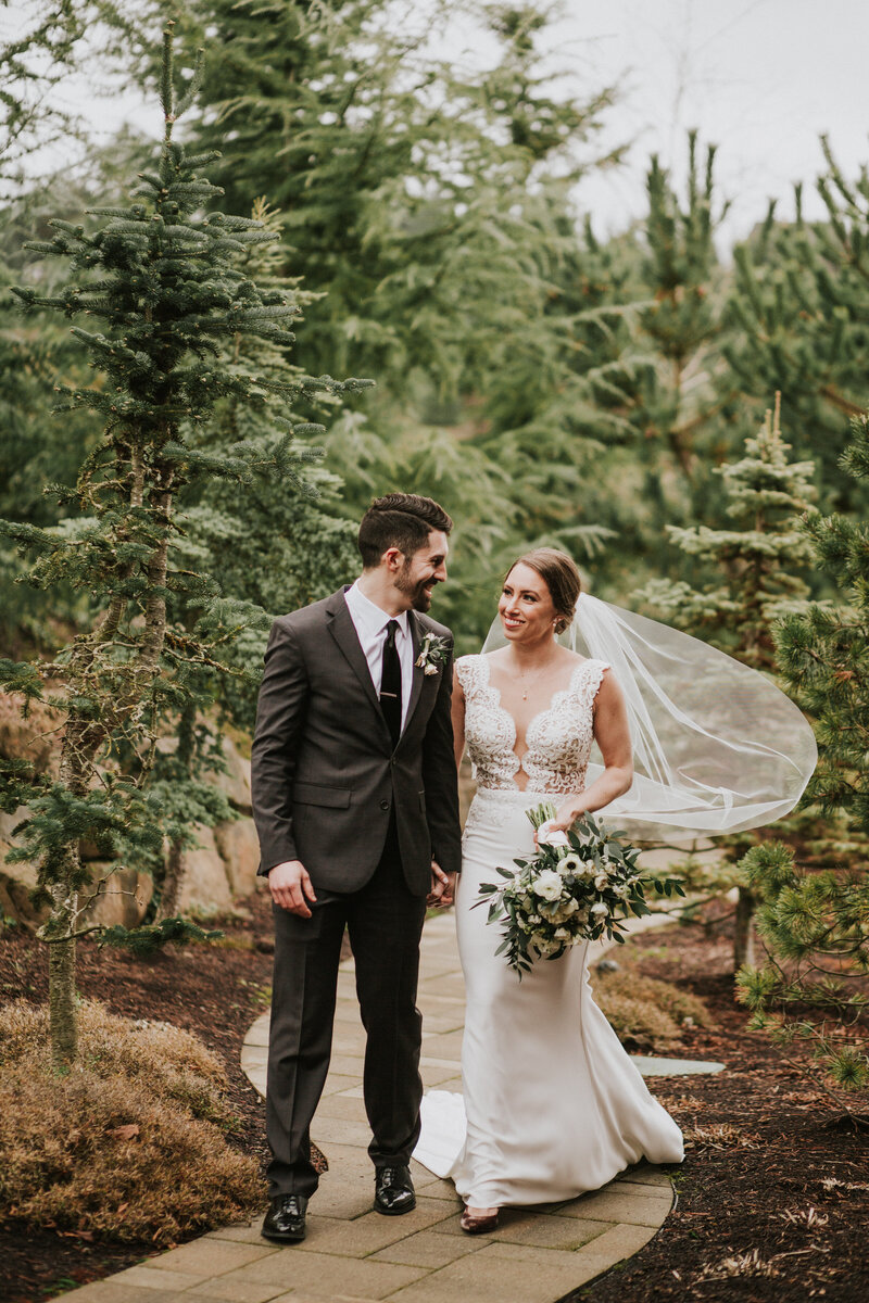 bride wearing a custom veil and holding a bouquet while walking down a path with her groom