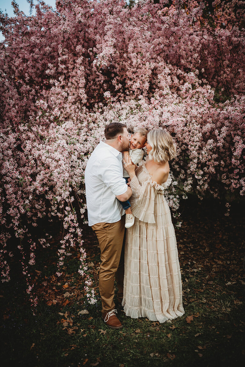 Family of three loving on each other in front of a beautiful blooming cherry tree.