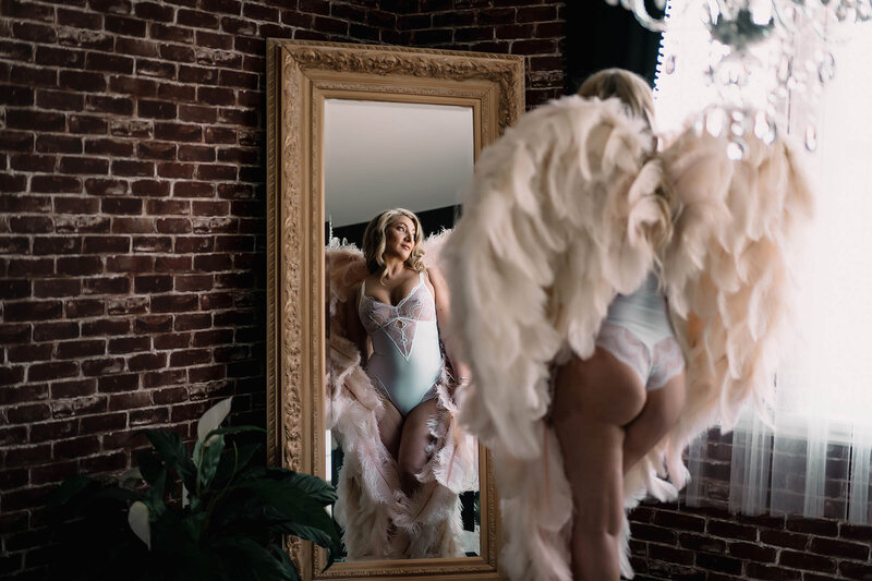 a woman in angel wings looks in the boudoir photoshoot mirror