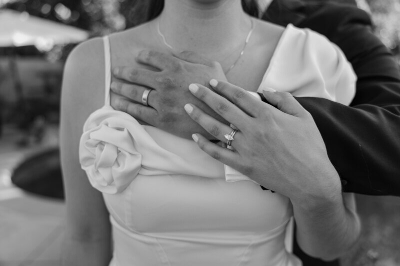 Details of the rings while the groom hugs the bride from behind and she rests her hand on his, in black and white.