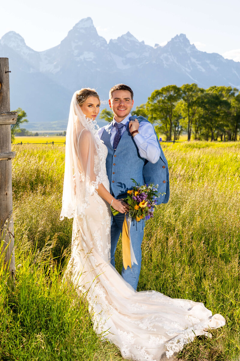 bride in a flowing lace gown and groom in a stylish blue vest stand in a sunlit Jackson Hole meadow, holding a vibrant wildflower bouquet, with the majestic Grand Teton peaks rising in the background for a luxurious wedding portrait.