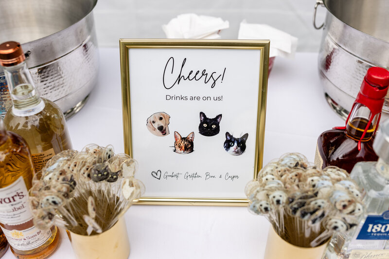 bar menu that says "cheers! drinks on us" with headshot photos of a white labrador retriever dog, calico cat, black cat, and  black and white cat on a table surrounded by liquor and drink stirrers in a cup with headshot photos of a white labrador retriever dog, calico cat, black cat, and  black and white cat 