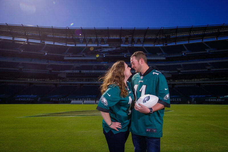 Engaged couple standing on Lincoln Financial Field in Eagles jerseys, holding a football and celebrating their shared passions.