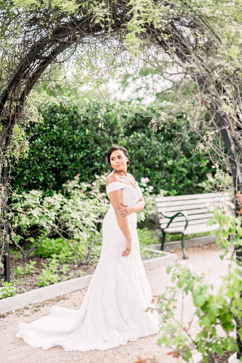 A bride looks over her shoulder, standing under an arch of branches in a green pathway.