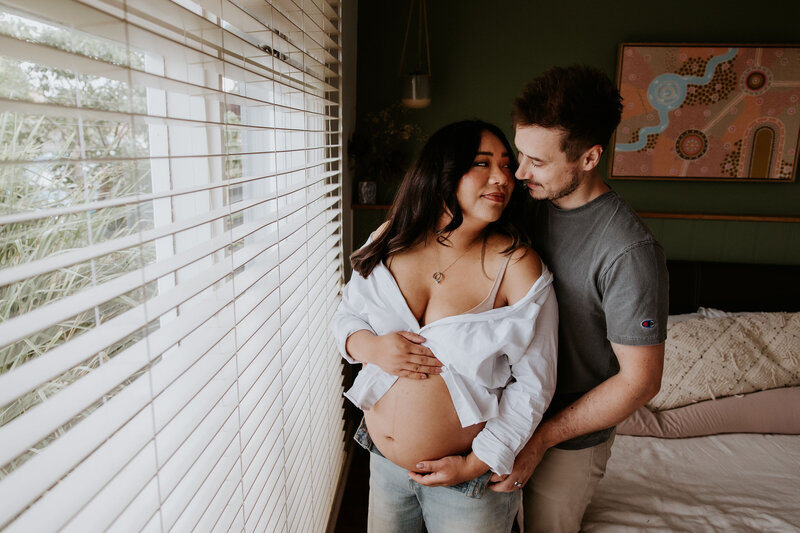 Man and pregnant woman cuddle at bedroom window