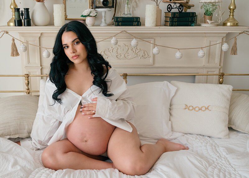 Mom poses in white button down on a bed with her hand on her pregnant belly.