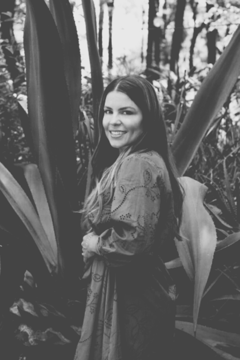 Natalie Bernacchi smiling in an agave garden, wearing a long green dress and standing among lush desert plants
