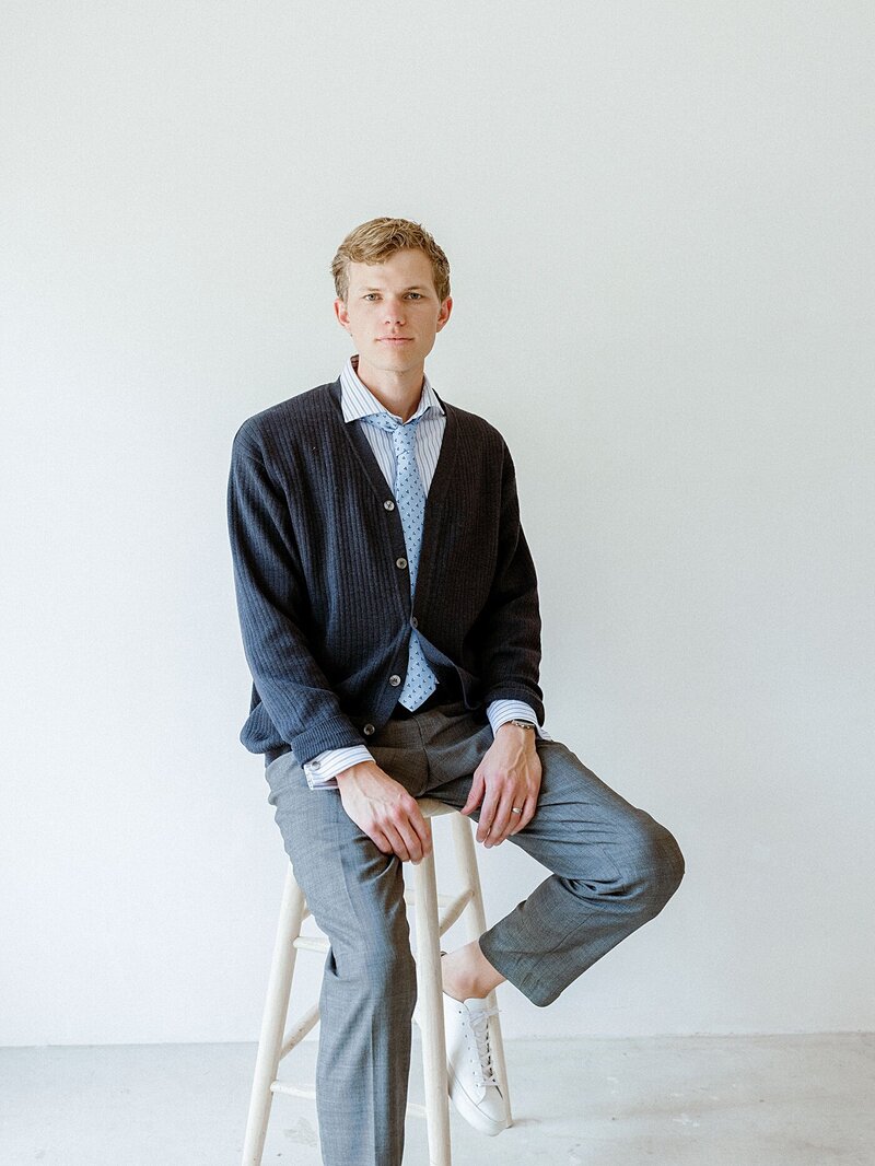 man sitting on a stool in a tie and sweater looking at the camera