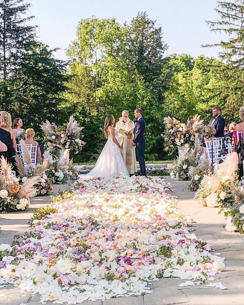 Bride and groom getting married outdoors with a priest, surrounded by beautiful flower petals laid in the aisle