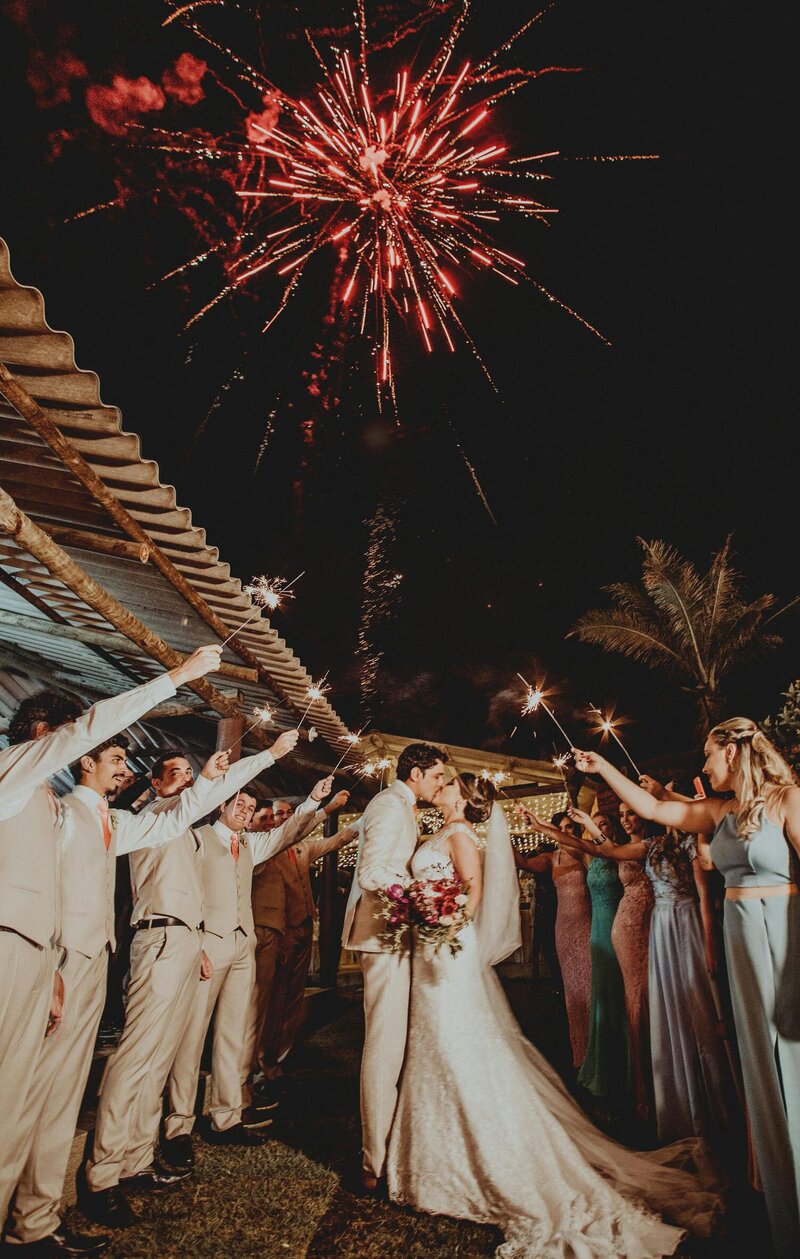 Exit photo of a bride and groom kissing under fireworks while the wedding party holds sparklers 