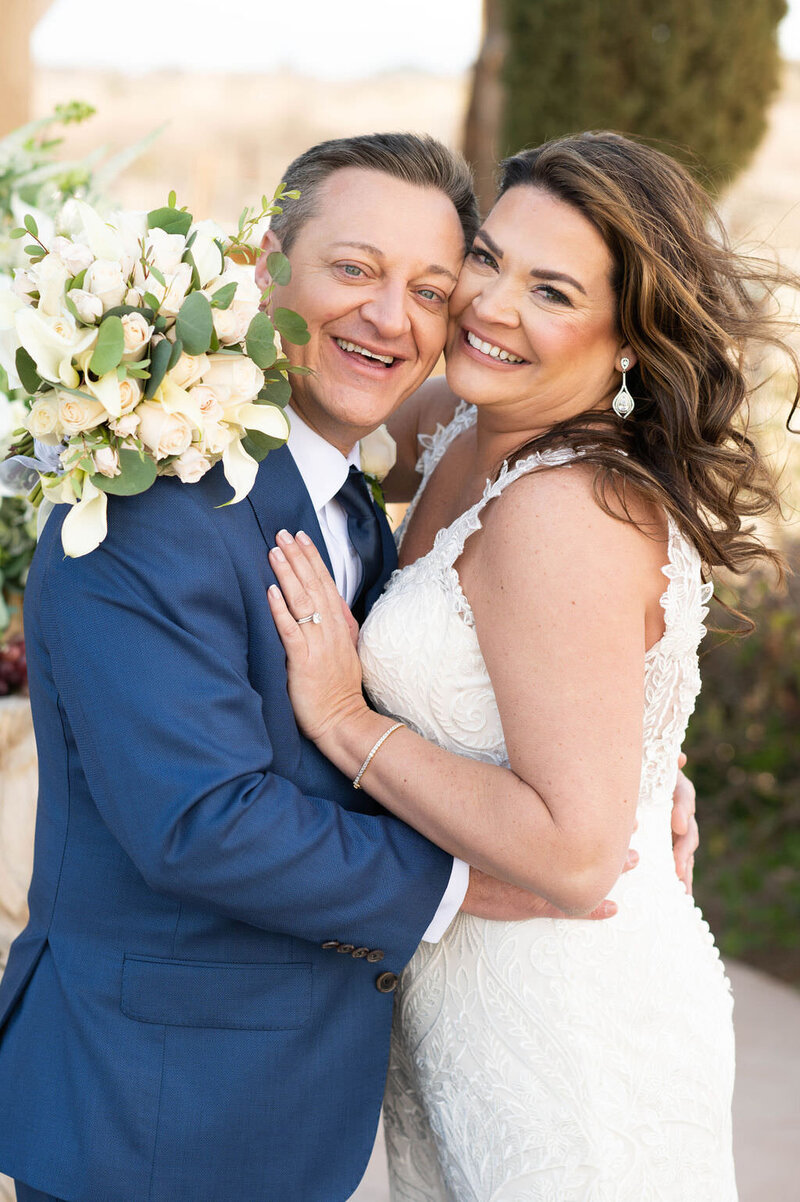 A newlywed couple smiling and hugging while one holds a bouquet of flowers.