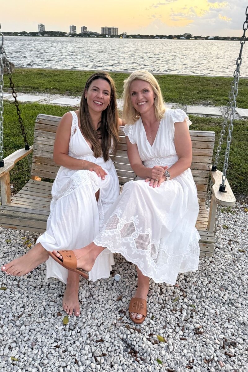 Natalie Bernacchi and Emily Collins smiling on a lakeside swing, connecting during their women’s mastermind retreat in white dresses