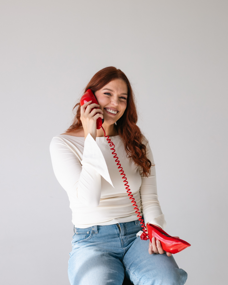 Mimi from Wed & Wilder smiling while talking on a playful red phone wearing a white top and jeans.