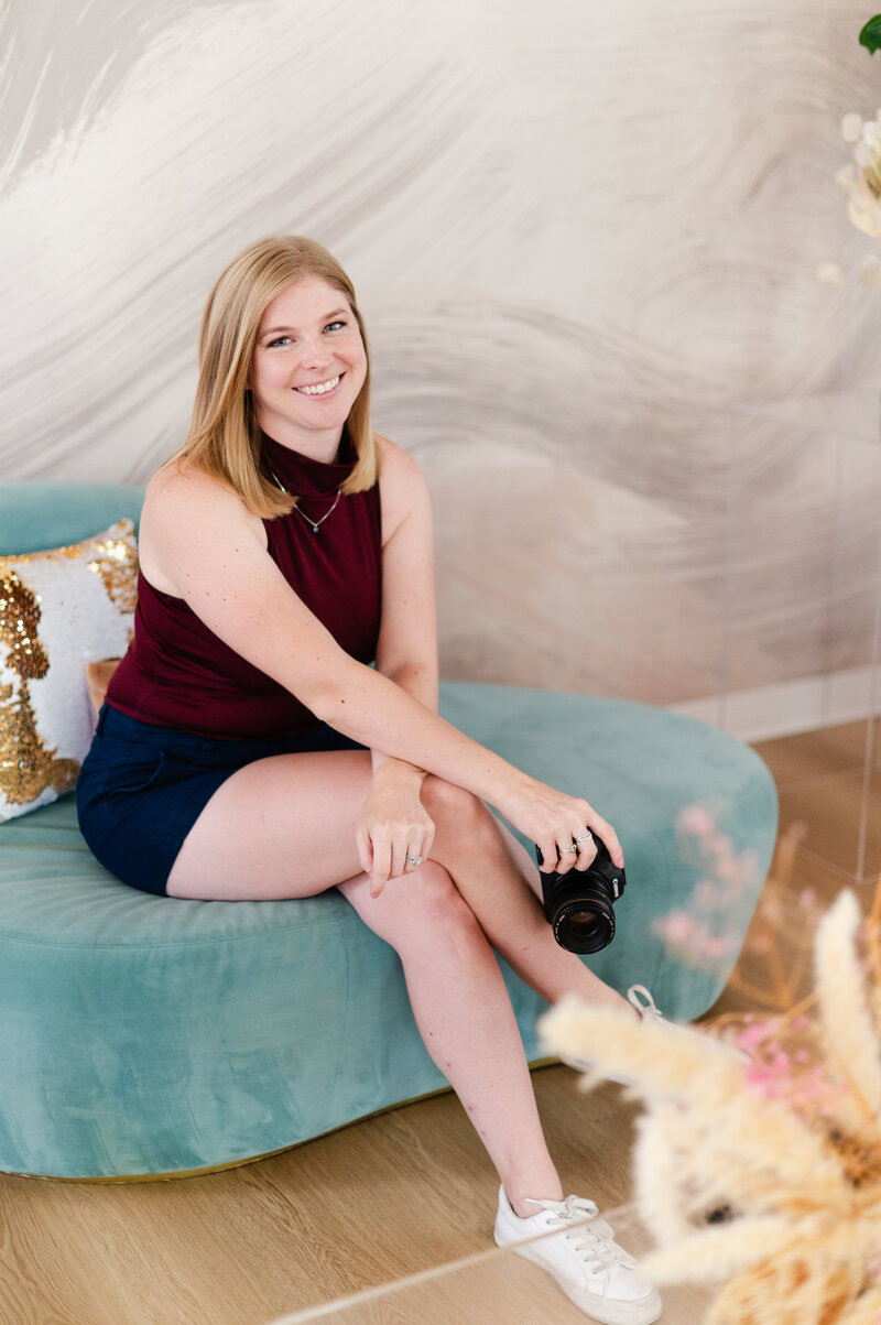 Image of Brook sitting on blue couch, smiling while holding her camera.