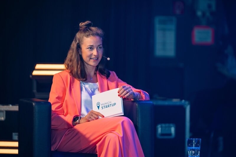 Moderatorin Hanna Zöller in rotem Anzug bei einer Live-Event Moderation für Munich Startup, mit Moderationskarte auf der Bühne
