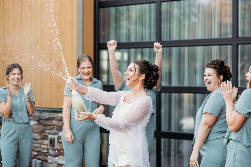 Photo of bride and bridesmaids in matching pajamas. Bride is popping champagne and bridesmaids are cheering for her. Photo by Ashley Swineford Photography