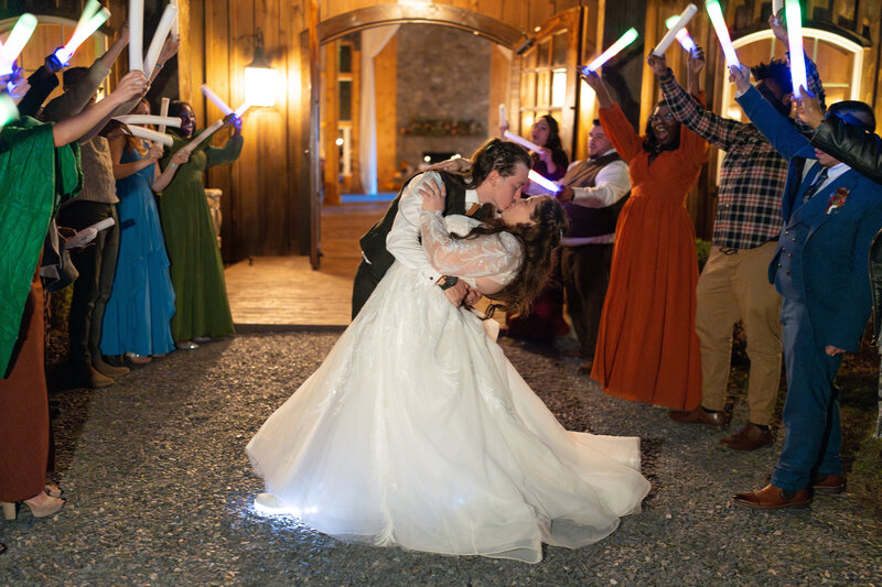 Bride and groom share a romantic dip kiss during their grand wedding exit at The Barn at Timber Creek in Farmville, VA, surrounded by guests waving glow sticks in celebration.