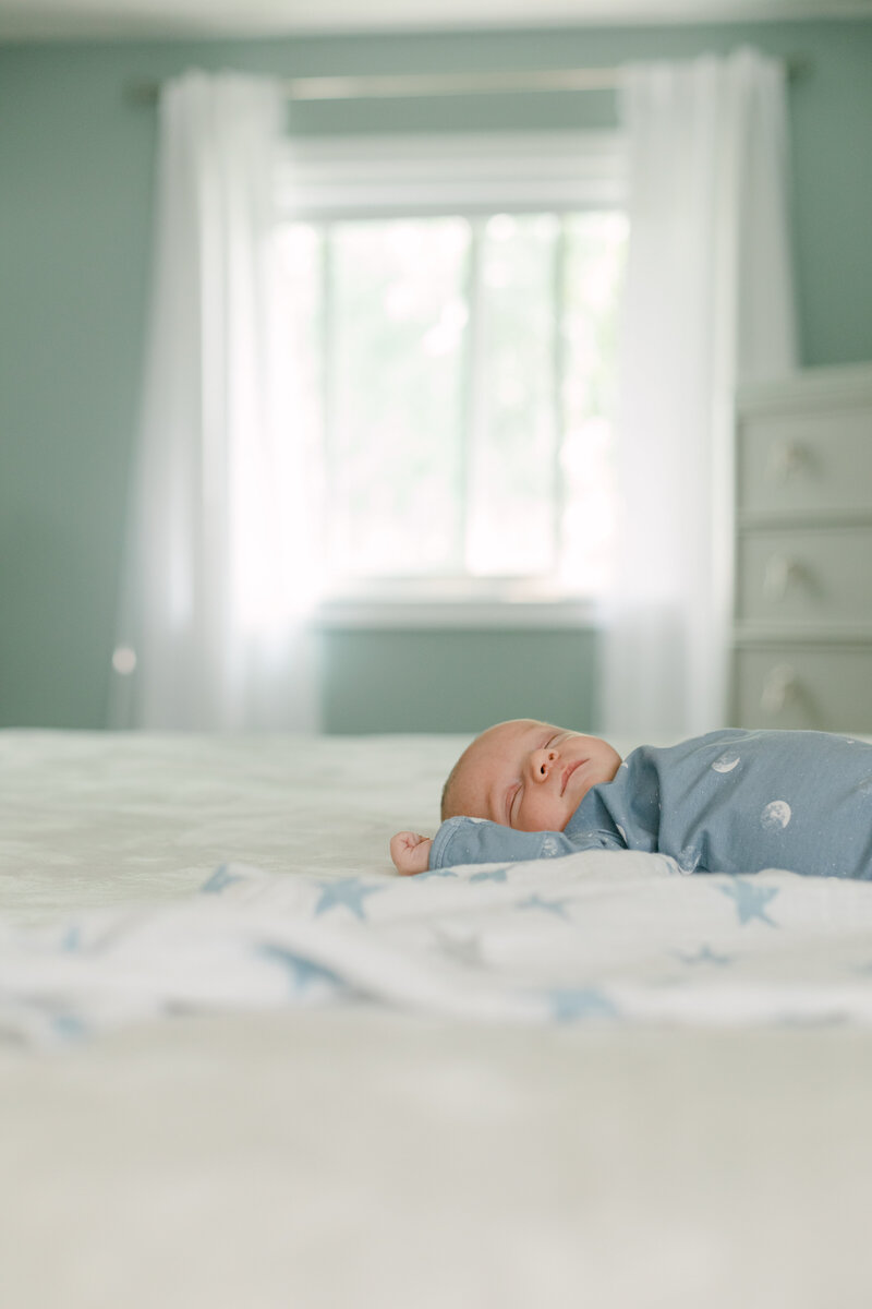 A newborn baby in a blue onesie sleeps peacefully on a bed with soft blankets. Natural light fills the cozy bedroom through a large window. Captured in Bowmanville, Ontario