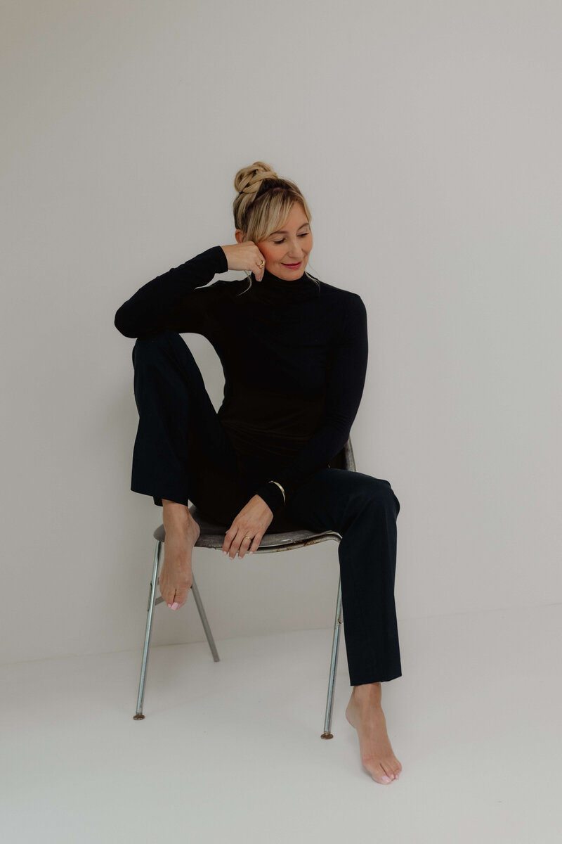 Women sitting barefoot in a chair with black turtle neck and black pants - resting her head in her hand.