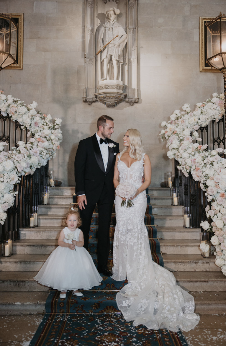 Danielle Armstrong and Tommy Edney at their UK wedding with daughter Orla, on a staircase adorned with flowers and candles.