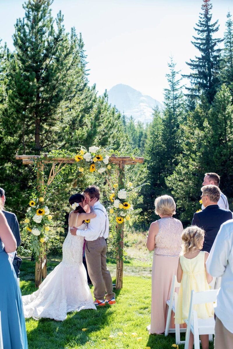 a man and woman kiss during their wedding ceremony with mt hood in the background and an arbor covered in sunflowers