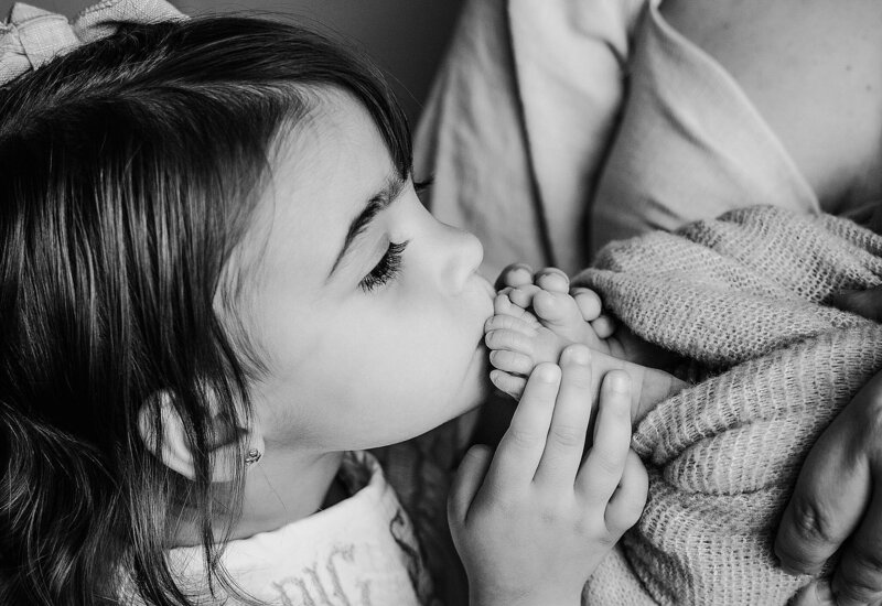A little girl kisses her mother's hand during a tender moment in a newborn session with her baby brother.