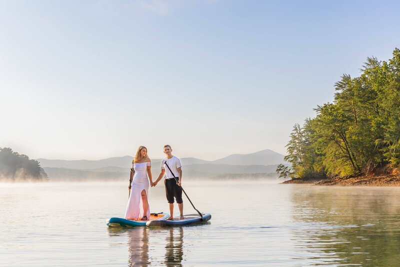A bride and groom stand on paddle boards in the middle of a lake. The bride is wearing a fitted strapless wedding gown and the groom is wearing a white t-shirt and rolled up black dress pants. There is a large mountain range behind them. The sun is shining as steam rises from the lake around them. The groom is smiling at the camera while the bride looks loving at the groom.