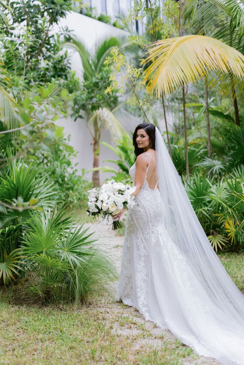Bride in a lace wedding gown with a long veil, holding a white bouquet and standing in a lush tropical garden on her destination wedding day