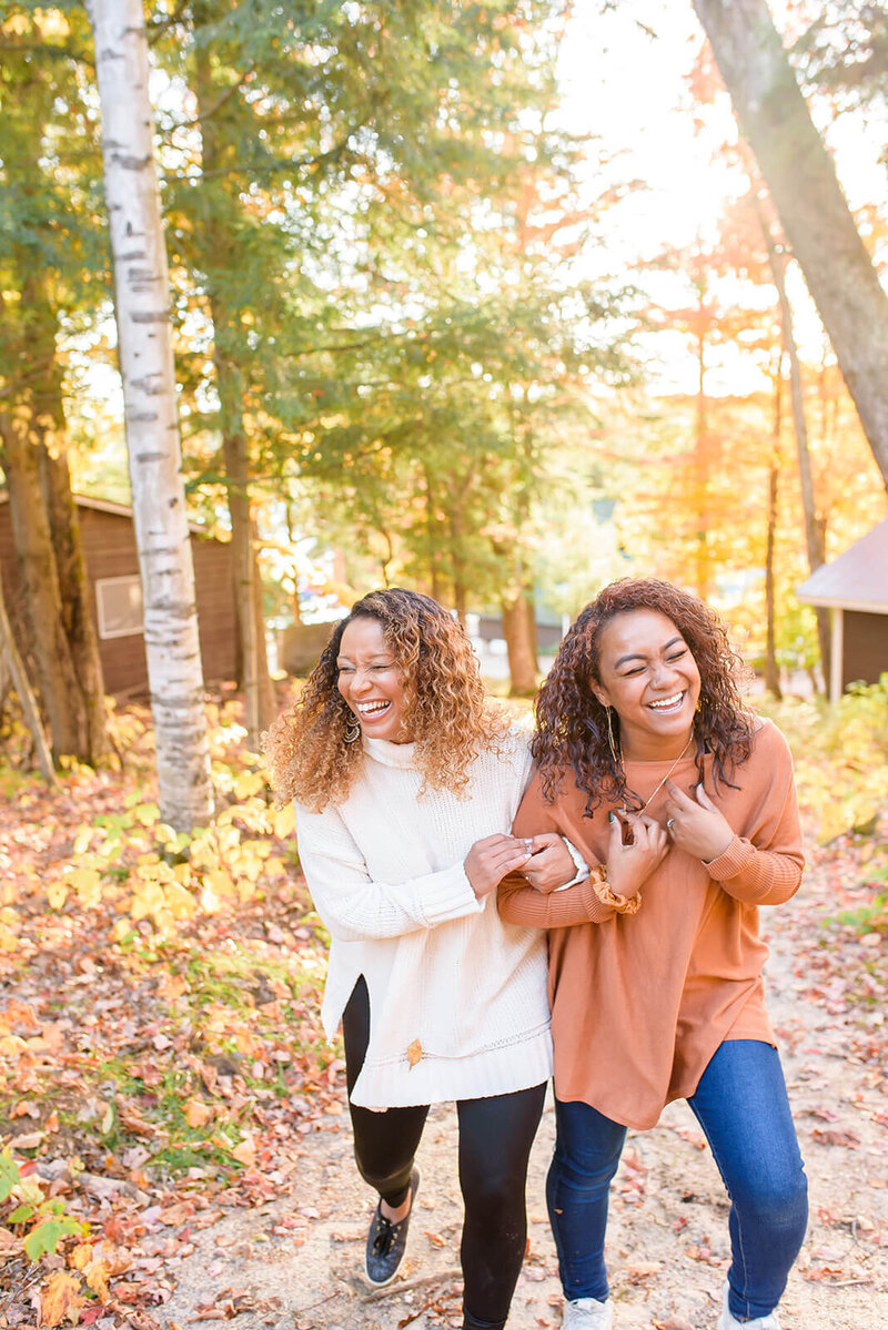 Two women. walking through a wooded path with cabins in the background. They are walking with arms linked and are laughing.