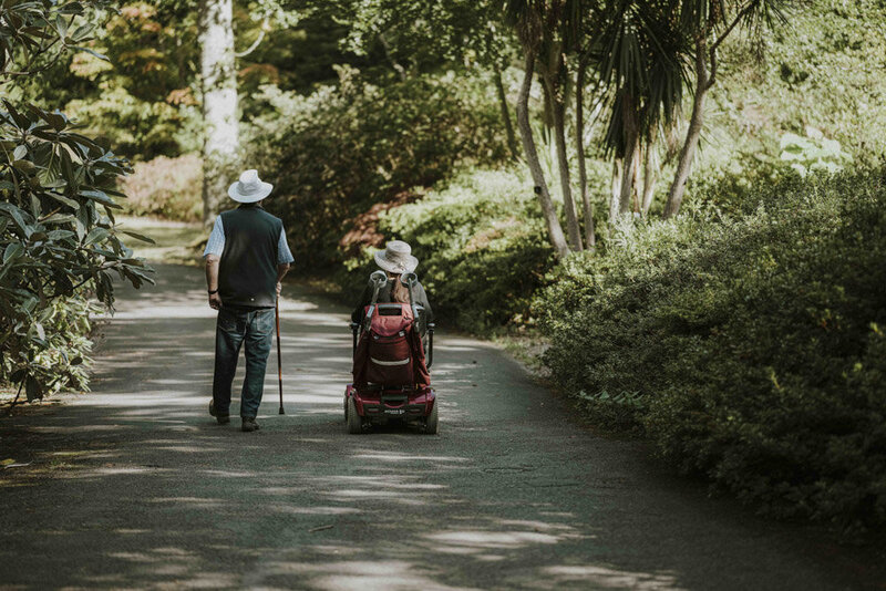 Two individuals are visible from the back, moving forward along a tree-lined, paved path. The partner on the left walks with a cane. The partner on the right sits in a red motorized wheelchair. They are both wearing sun hats.