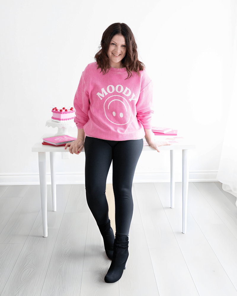 Emily, brand and web designer, standing in front of computer desk in a pink sweatshirt.