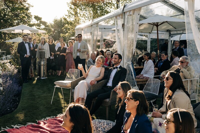 wedding guests listen to speeches while sitting outside in the sunset on white wire chairs from little hire co