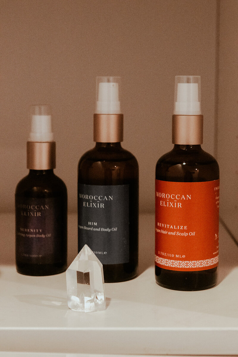 Three Moroccan Elixir sprays (Revitalize, him and serenity) sit next to each other with quartz crystal  sitting in front of them