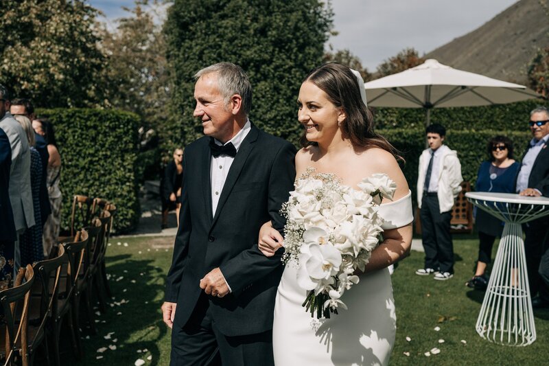 father walks bride carrying white bouquet down aisle in summer at winehouse queenstown